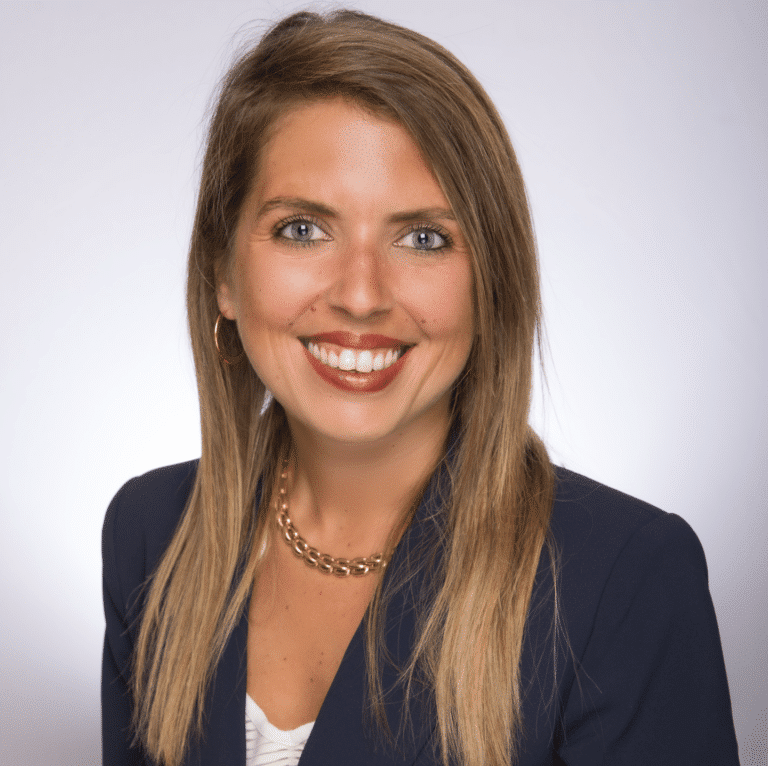 A woman with long blonde hair, wearing a navy blazer, white top, gold chain necklace, and hoop earrings, smiles at the camera against a plain light background.